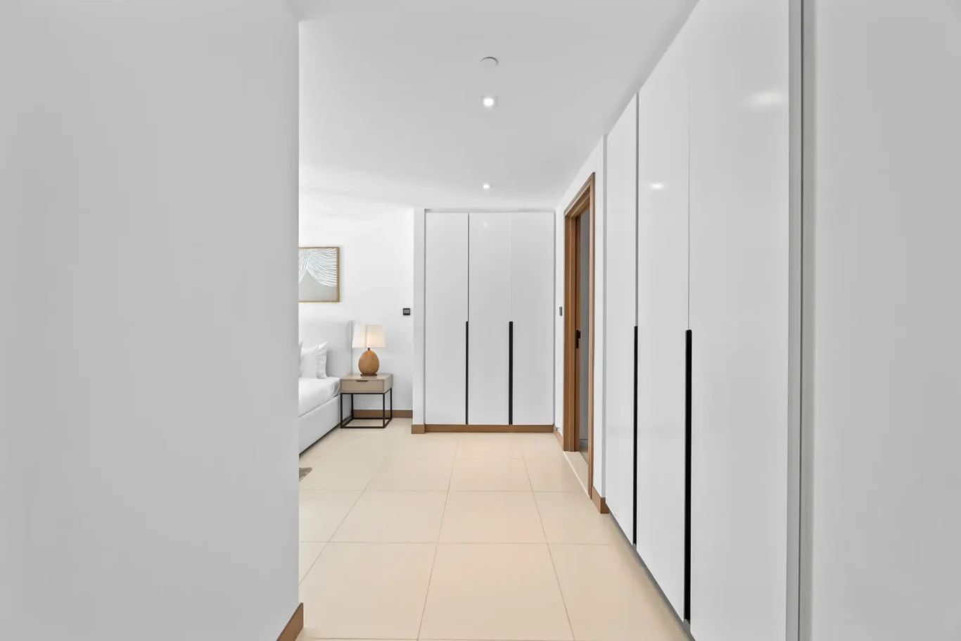 Bright hallway with white built-in wardrobes and a bed visible at the far end; warm bedside lamp on a small table beside the bed.