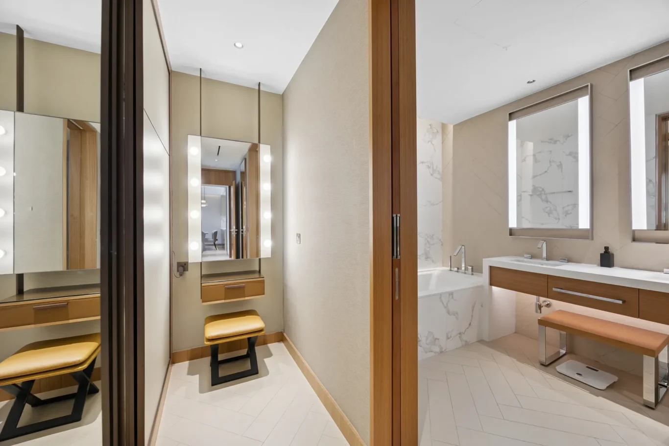 Modern en-suite view: marble walls, backlit mirrors, and a wooden vanity with stools in a dressing area adjacent to a bathtub area.