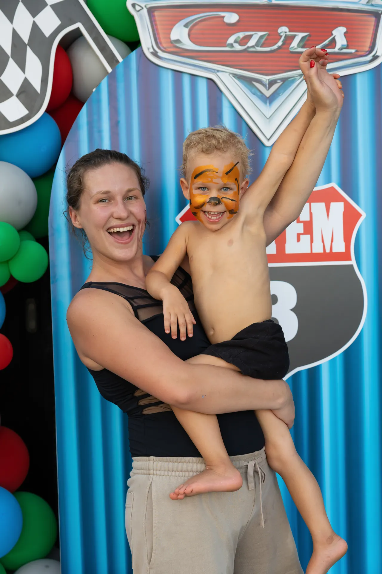 Smiling woman in a black top lifts a shirtless child with tiger face paint against a blue race-themed backdrop and balloons behind them.
