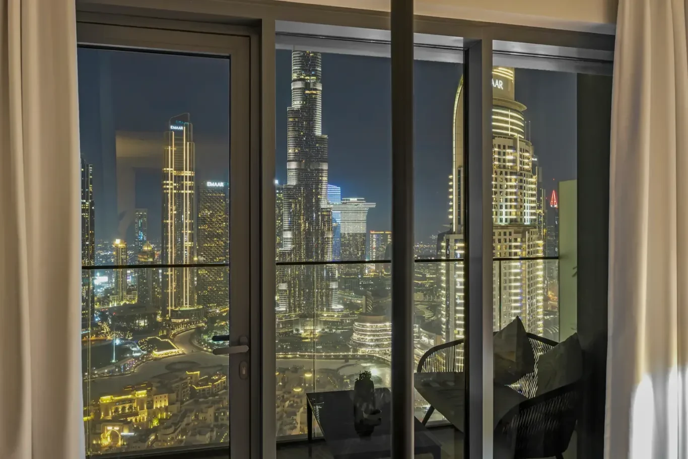 Night city skyline through large glass doors from a high-rise home; illuminated skyscrapers outside, with a small table inside.
