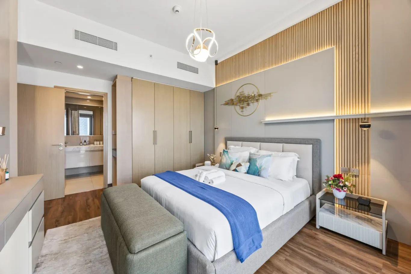 Modern hotel bedroom with a king bed, gray upholstered headboard, blue throw, and a warm wood accent wall with subtle lighting.