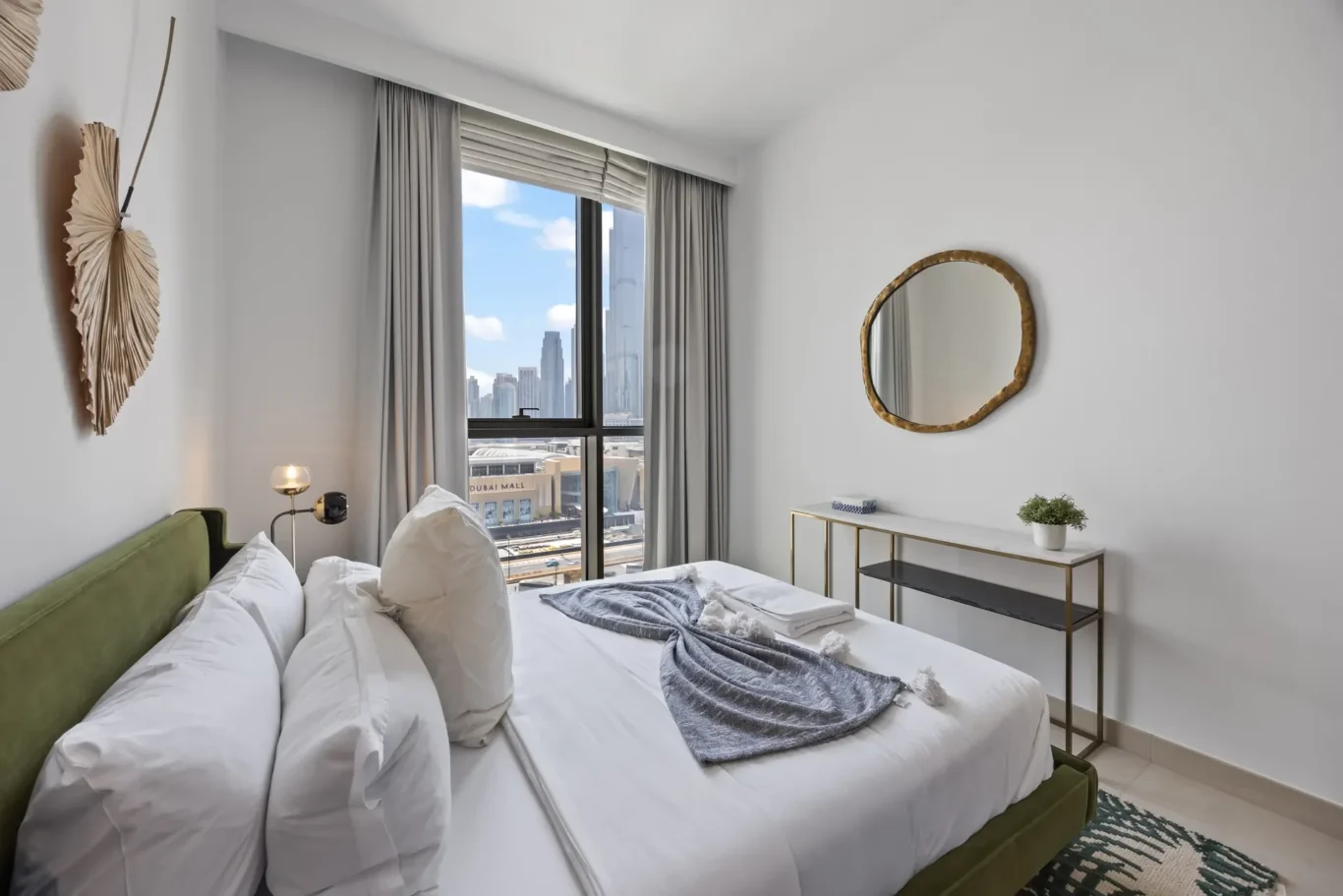 Hotel bedroom with a large window showing a city skyline and blue sky; white bedding, green headboard, and a round gold-framed mirror on the wall.