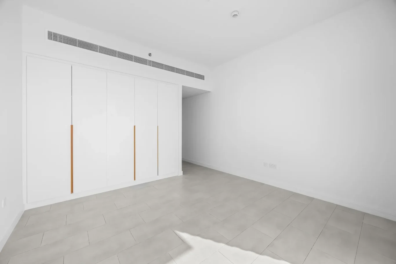 White built-in wardrobes with gold vertical handles along a wall in a minimalist empty room.