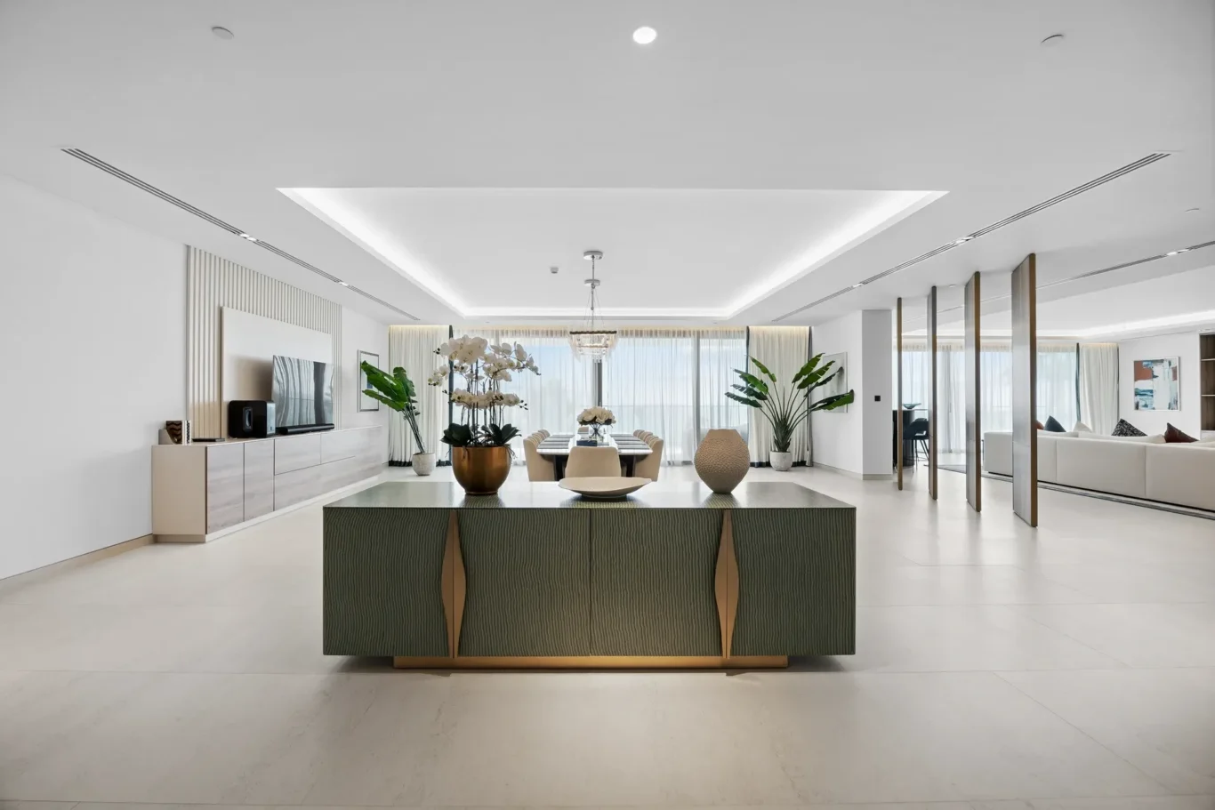 Bright, open-concept living and dining area with a central dark green cabinet featuring gold accents and large potted plants.