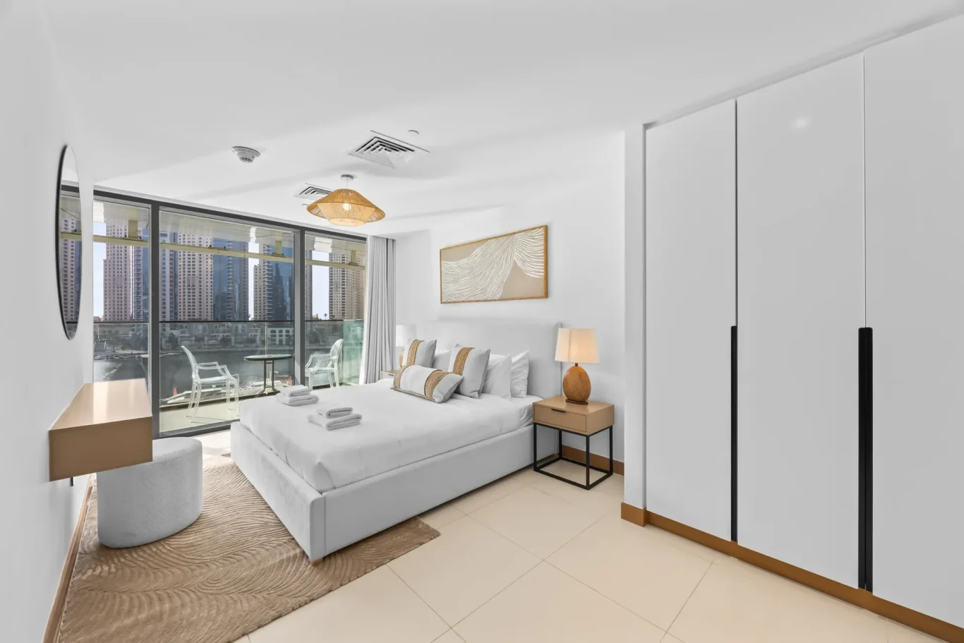 Modern hotel bedroom with a large bed, white linens, and a balcony view of city skyscrapers through glass doors.