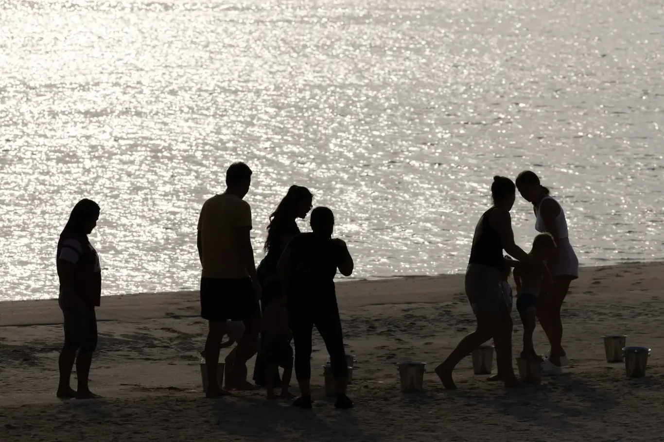Silhouetted group of adults and children on a sandy beach near the glistening water, with several buckets nearby.