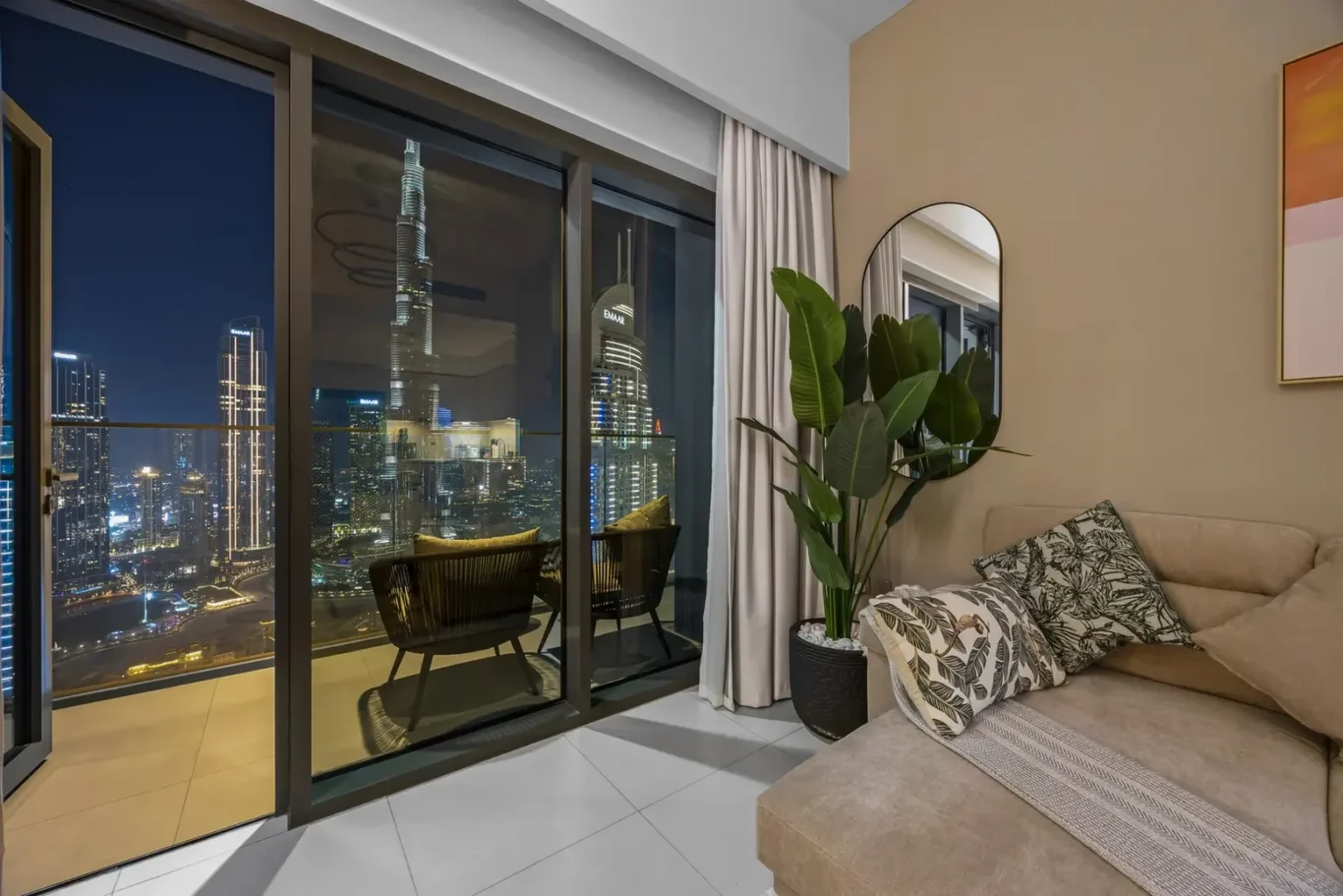 Night view of a city skyline from a modern living room with a balcony and beige sofa in the foreground.