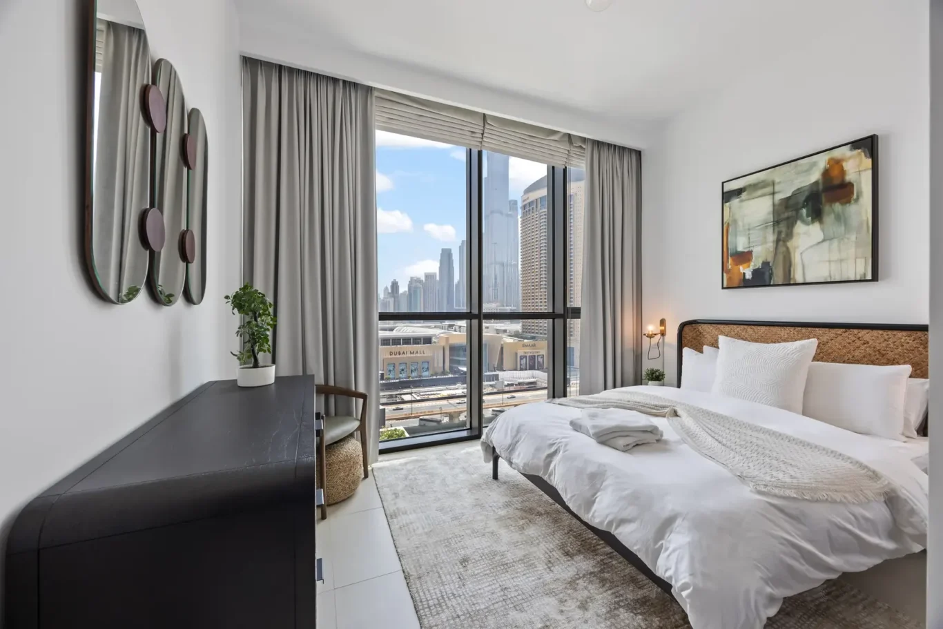 Bright hotel bedroom with a large window overlooking the Dubai skyline, king bed, abstract artwork, and dark wood dresser on the left.