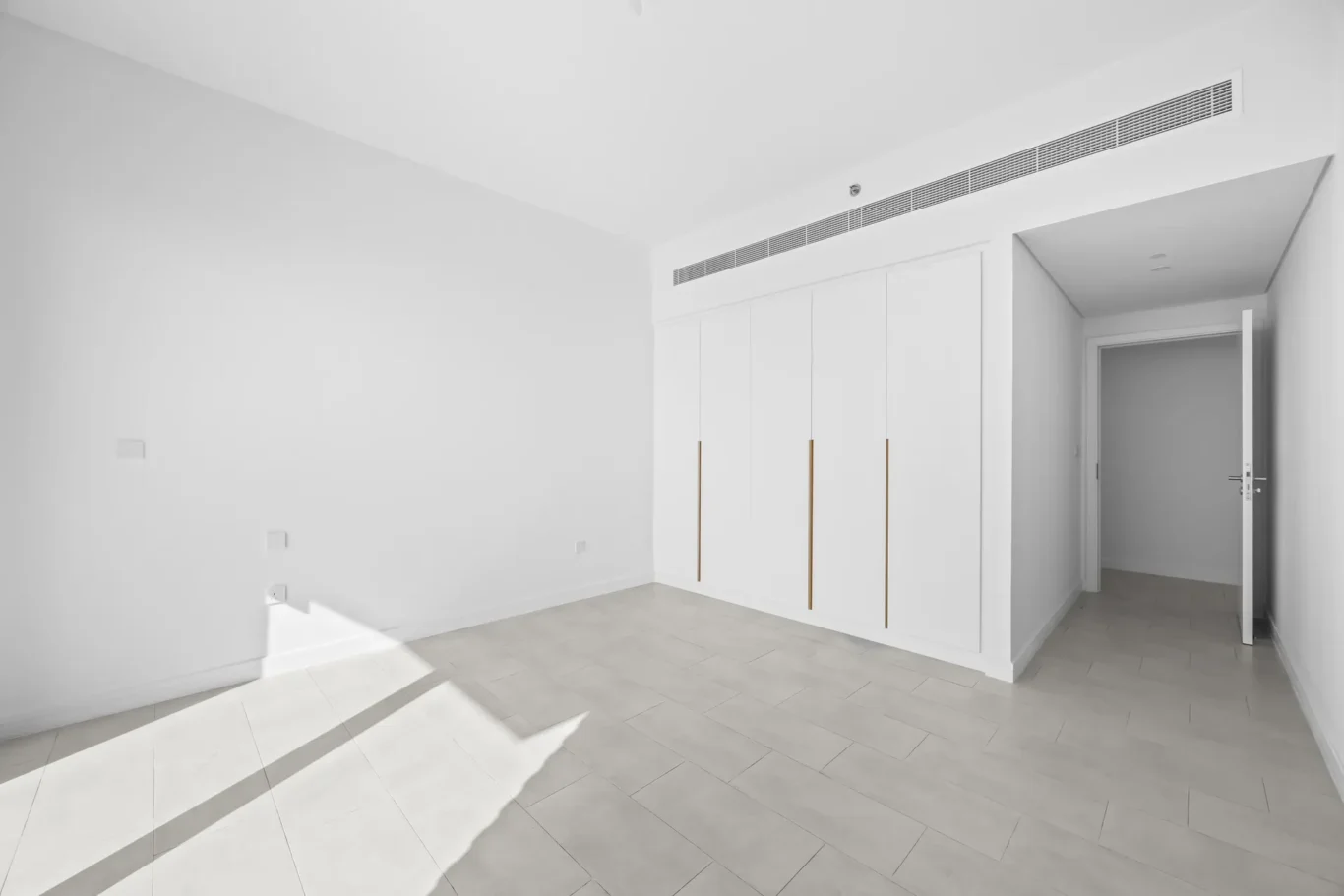 Bright, empty white room with built-in white closets and a long ceiling vent above; doorway open to a hallway/room beyond the closets.
