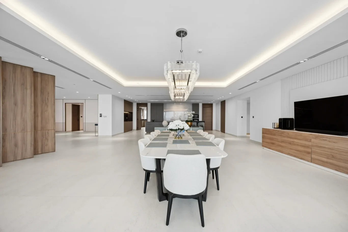 Bright open-concept dining area with a long white table, white chairs, and a crystal chandelier centerpiece.