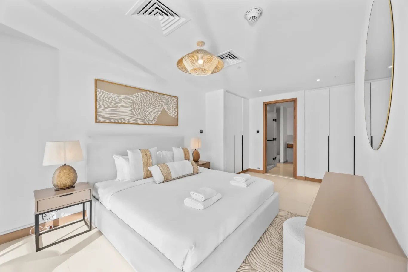 Bright, minimalist hotel bedroom with a large neatly made bed, wicker bedside tables and lamps, white wardrobes, and a round wall mirror on the right; doorway to an en-suite visible in the background.