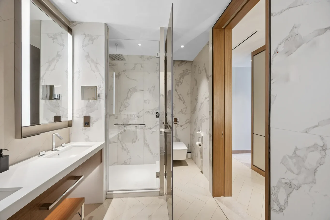 Modern marble bathroom with a glass shower enclosure, double sink vanity, and wood accents on the doorway. It’s a bright, contemporary design.