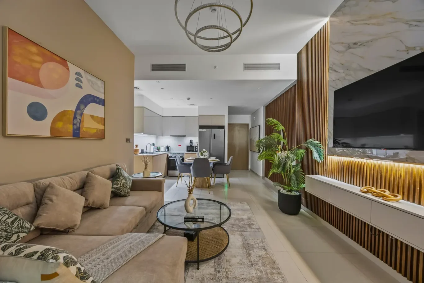 Open-plan living room and kitchen with a beige sectional, round glass coffee table, abstract wall art, and a wooden slat wall beside a large TV.