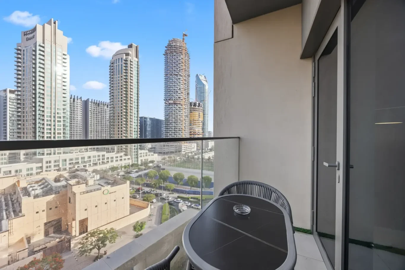 Balcony with a round black table and two chairs overlooking a city skyline of tall modern skyscrapers, one under construction, under a blue sky.