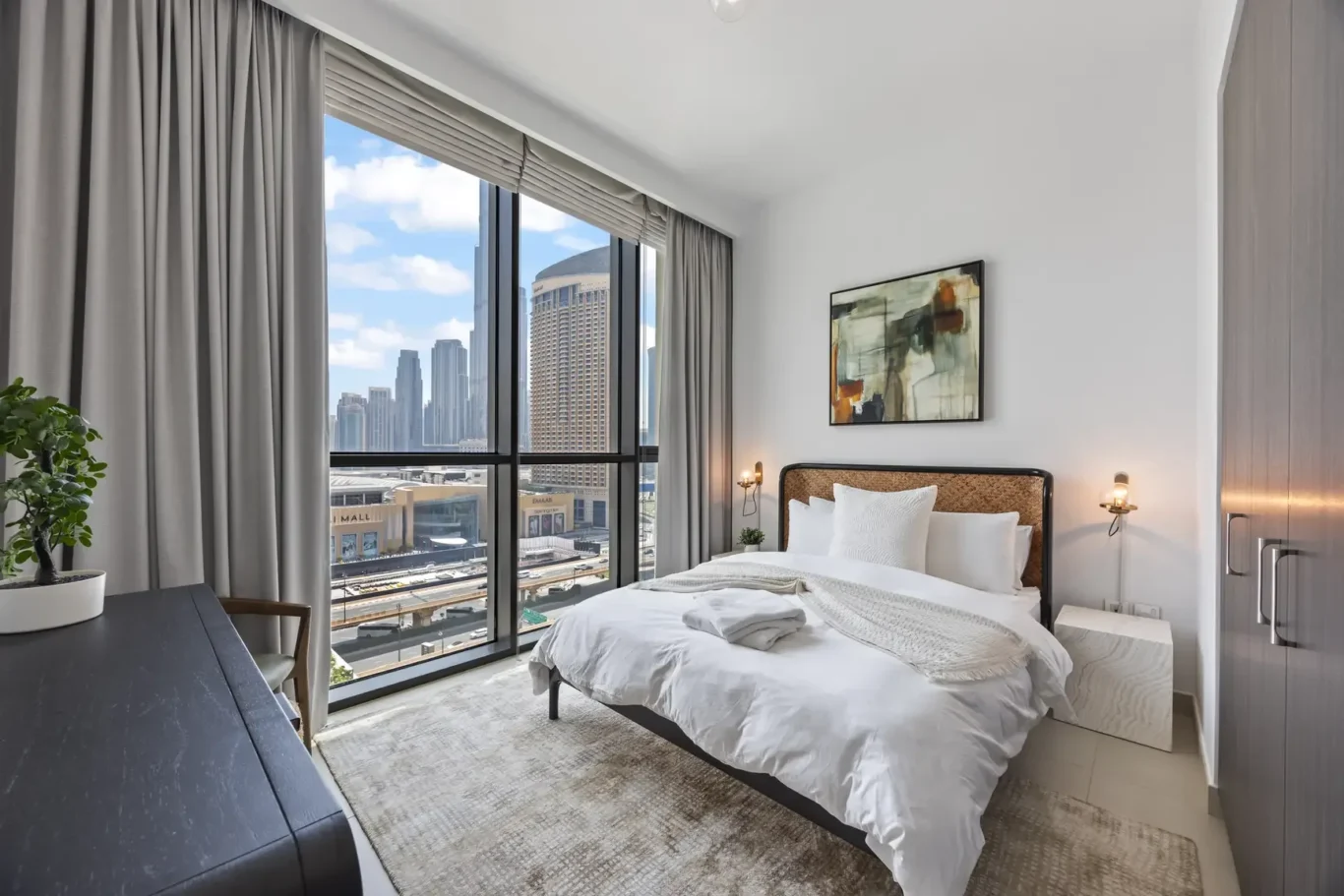 Modern hotel-style bedroom with a large window showing a city skyline, a white made bed, and abstract wall art.