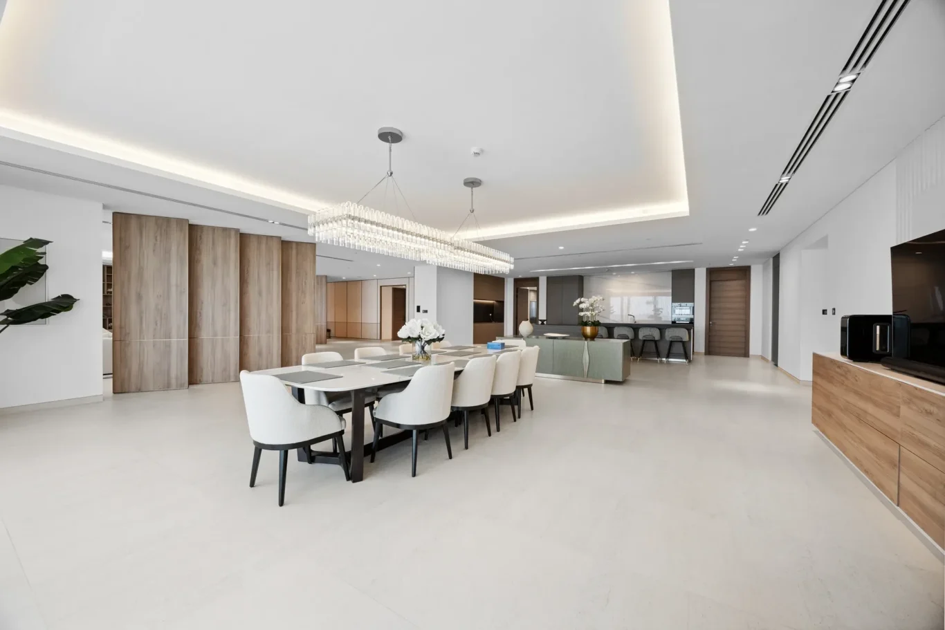 Open dining area with a long white table, white upholstered chairs, and a crystal chandelier; modern kitchen in the background.