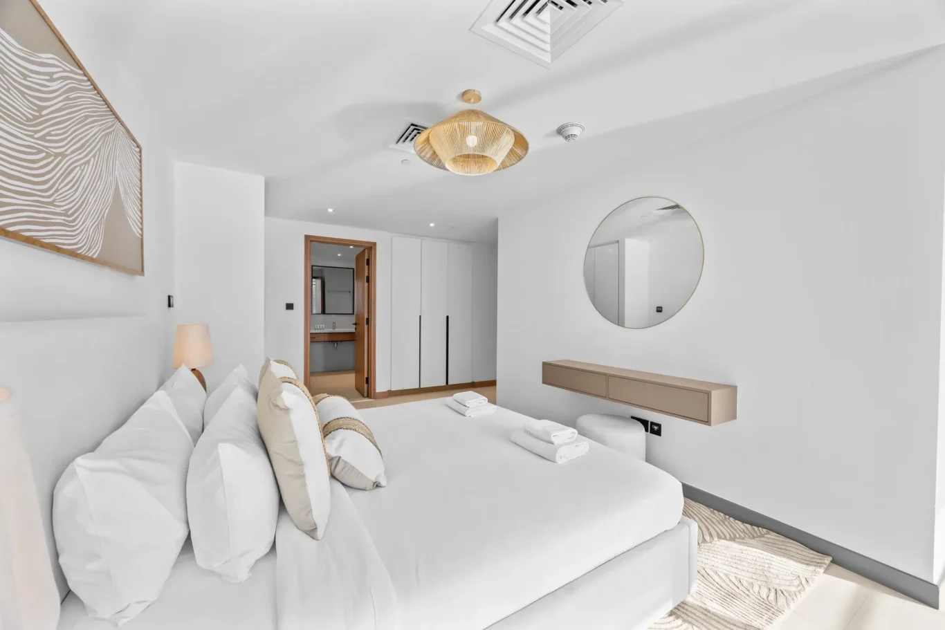 Minimalist white hotel bedroom with a large bed, decorative pillows, and a round wall mirror opposite the bed.