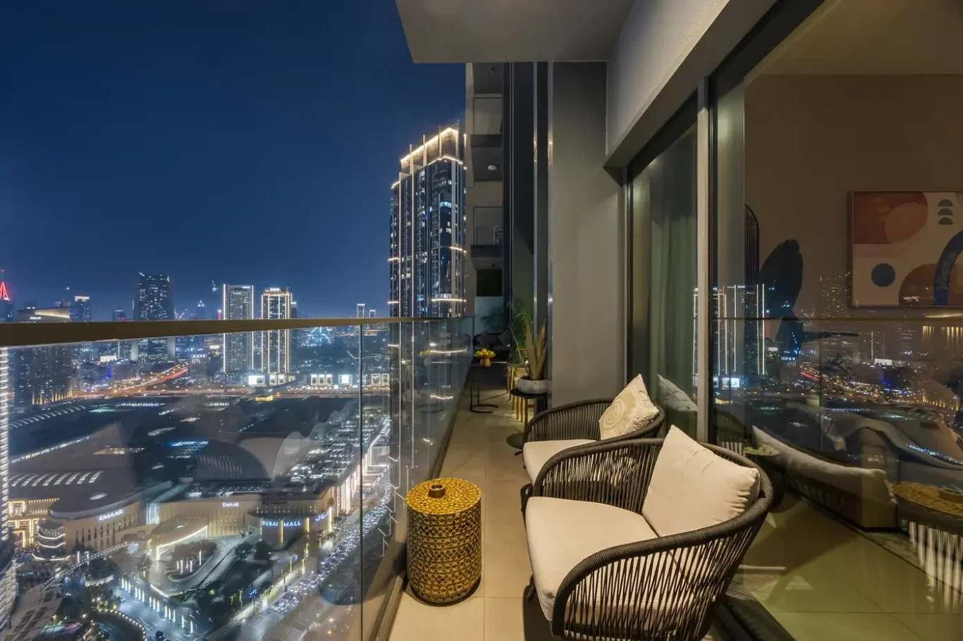 Night view from a high-rise balcony with glass railing, wicker chairs and a yellow woven table overlooking a brightly lit city skyline at night,