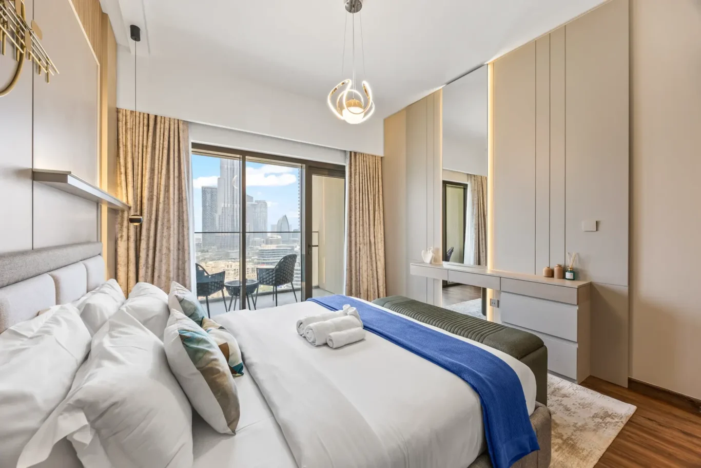 Luxurious hotel bedroom with white linens and a blue throw, facing a balcony with a city skyline view.