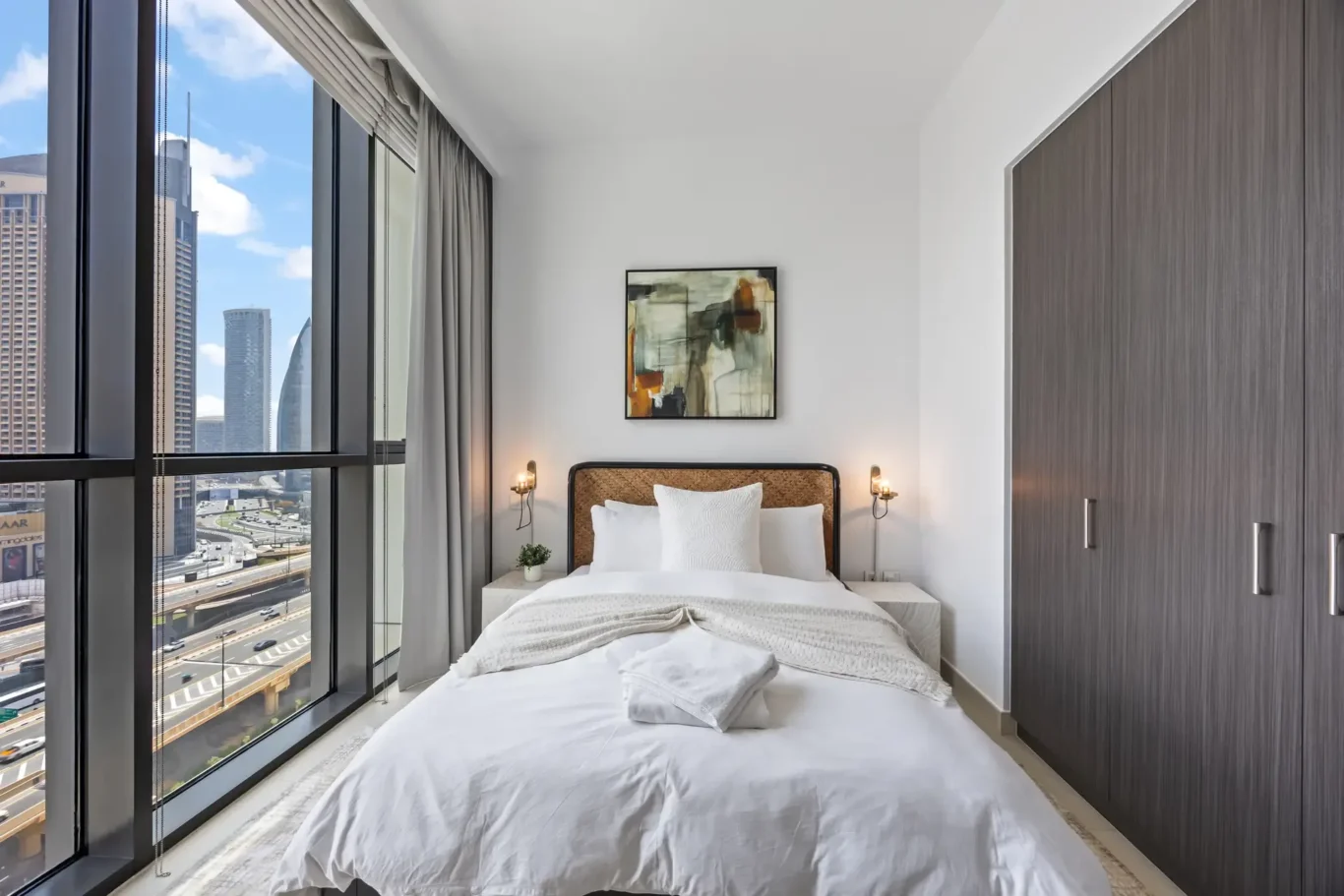 Modern hotel bedroom with floor-to-ceiling windows showing a city skyline, white bedding, and abstract art above the bed.