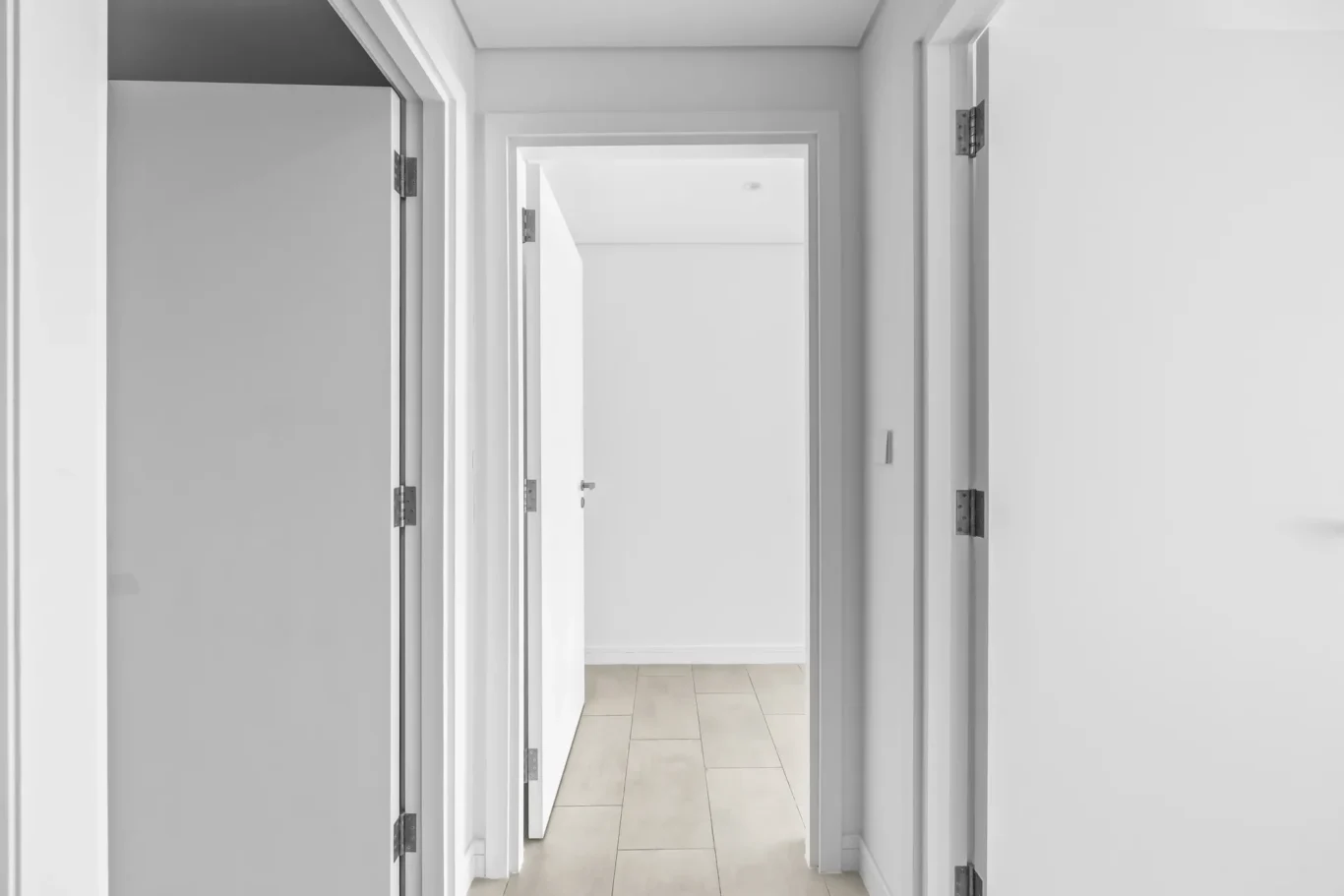White hallway with multiple white doors on both sides and light tiled floor, leading to a bright room ahead.