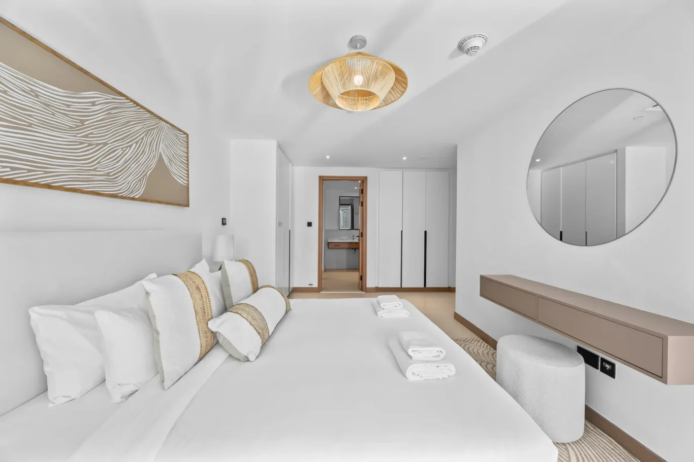 Bright hotel-style bedroom with a large white bed, wicker pillows, and a tan-framed wave-pattern wall art.