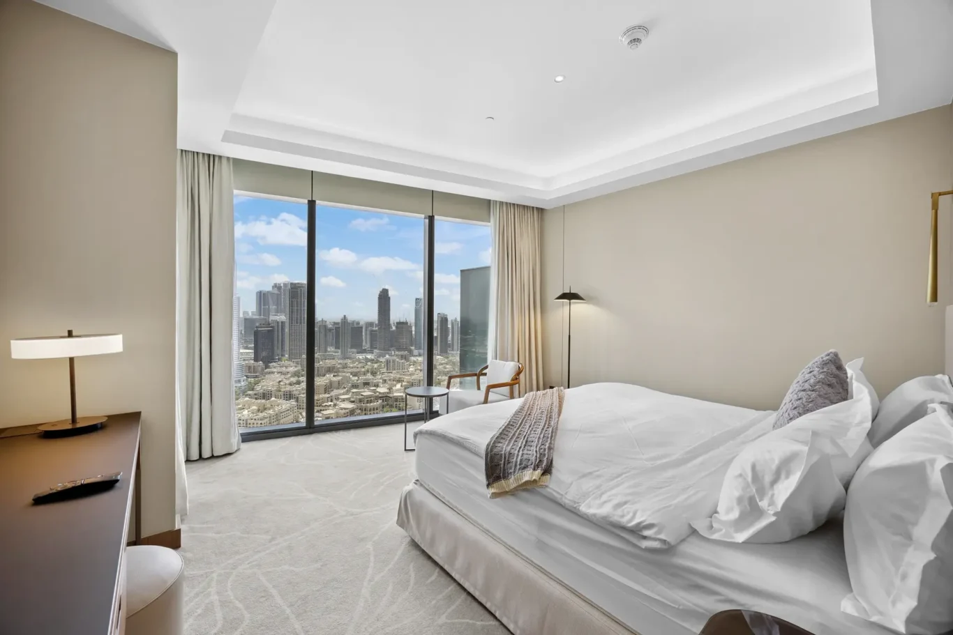 Hotel bedroom with two white beds, beige walls, and a floor-to-ceiling window showing a city skyline.