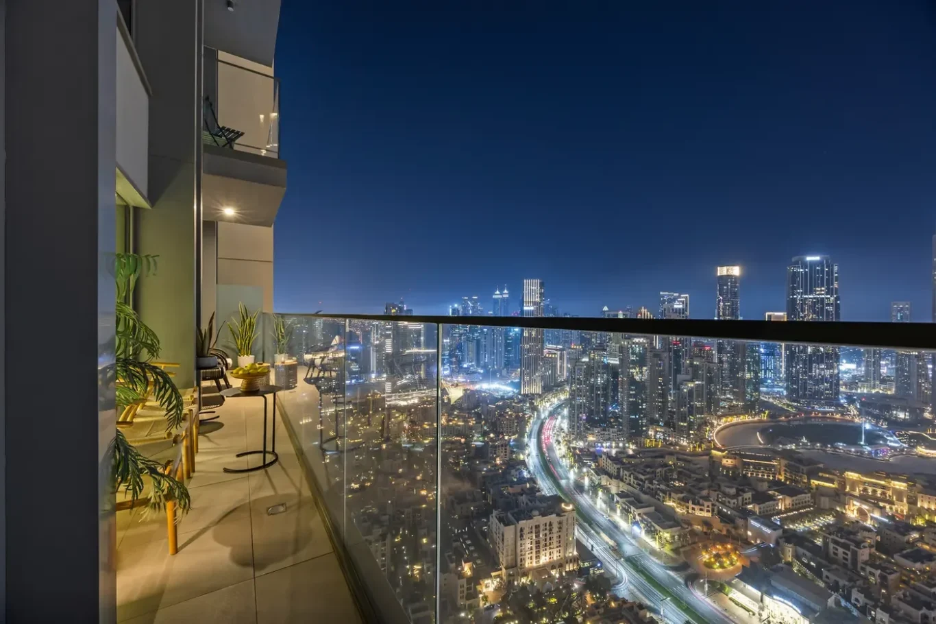 Balcony at night with glass railing overlooking a brightly lit city skyline and a seating area with plants.