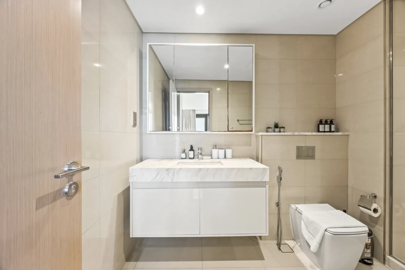 Modern bathroom with a marble countertop sink, wall mirror, and beige tiled walls. A white floating vanity sits under the mirror.