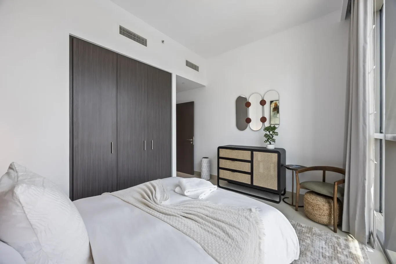 Bedroom with white walls, a dark wood wardrobe on the left, and a cane-drawer dresser with a plant on top.
