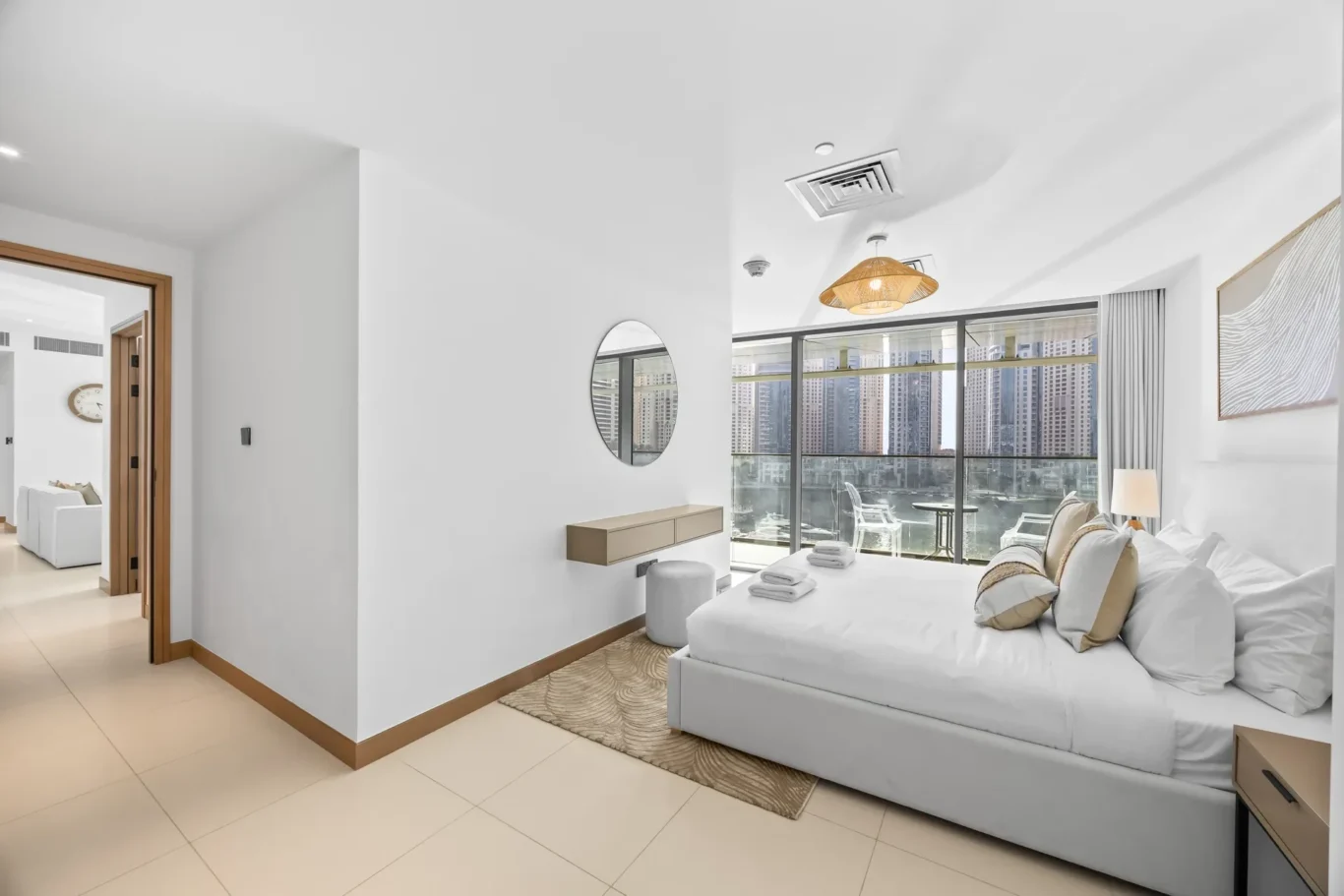 Bright hotel bedroom with large glass balcony doors, white bedding, and a city skyline view outside.