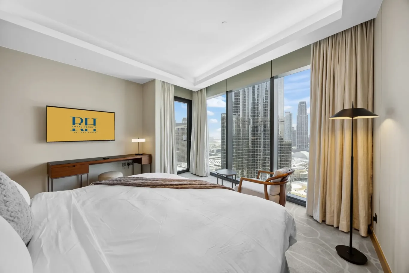 Modern hotel room with two white beds, a desk and a wall-mounted TV, and floor-to-ceiling windows showing a city skyline.