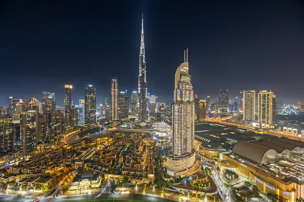 Nighttime city skyline of Dubai with the Burj Khalifa and illuminated skyscrapers under a dark sky, seen from above.