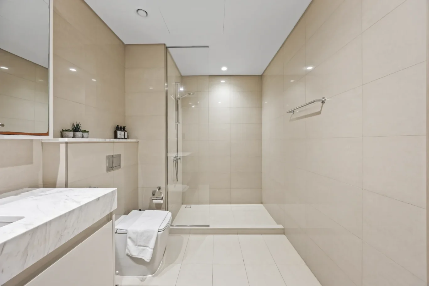 Bright modern bathroom with glass shower enclosure, beige tiled walls, and marble countertop.