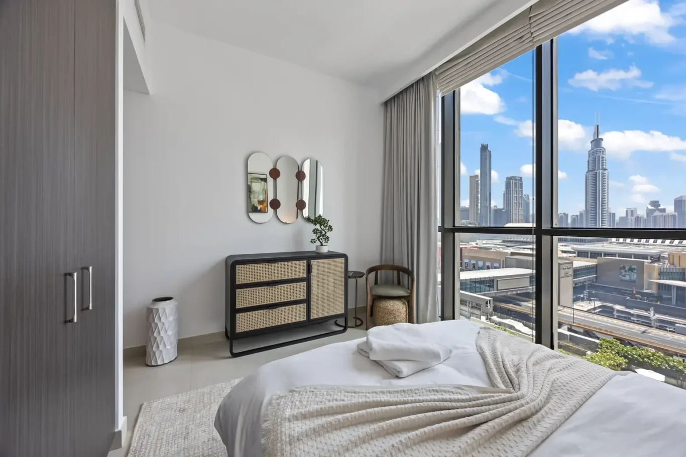 Bright modern bedroom with a large window view of a city skyline, white bedding, and a black dresser with woven panels.