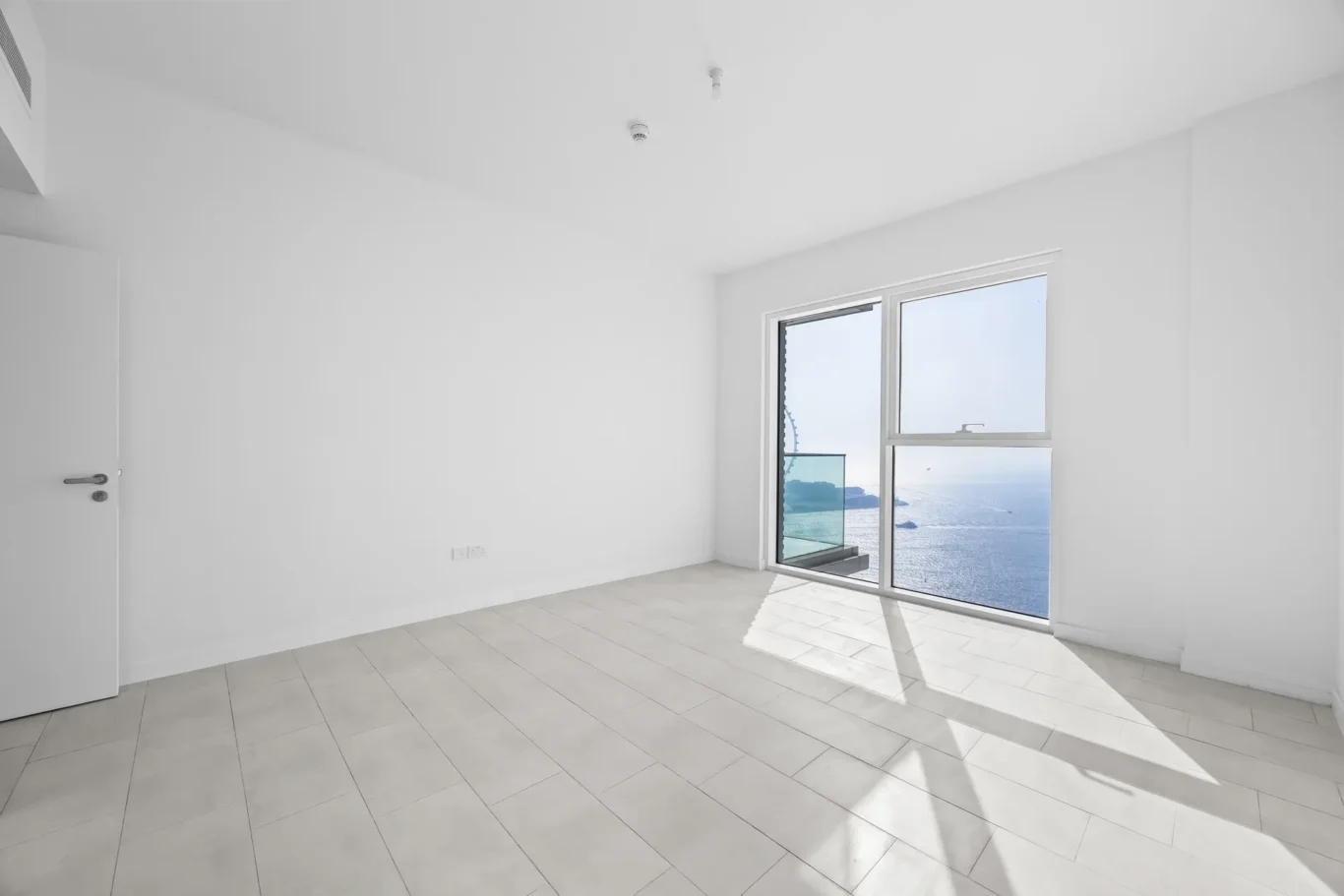 Empty bright room with a large glass door/window showing the ocean, white walls and tiled floor, left doorway present.