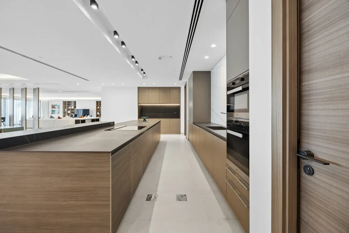 Modern kitchen with a long wooden island, beige cabinetry, and integrated ovens, open to a bright living area.