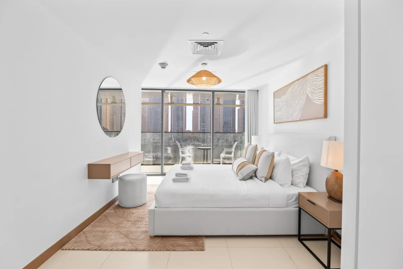 Bright hotel-style bedroom with white bedding, beige rug, and a city skyline visible through glass balcony doors.