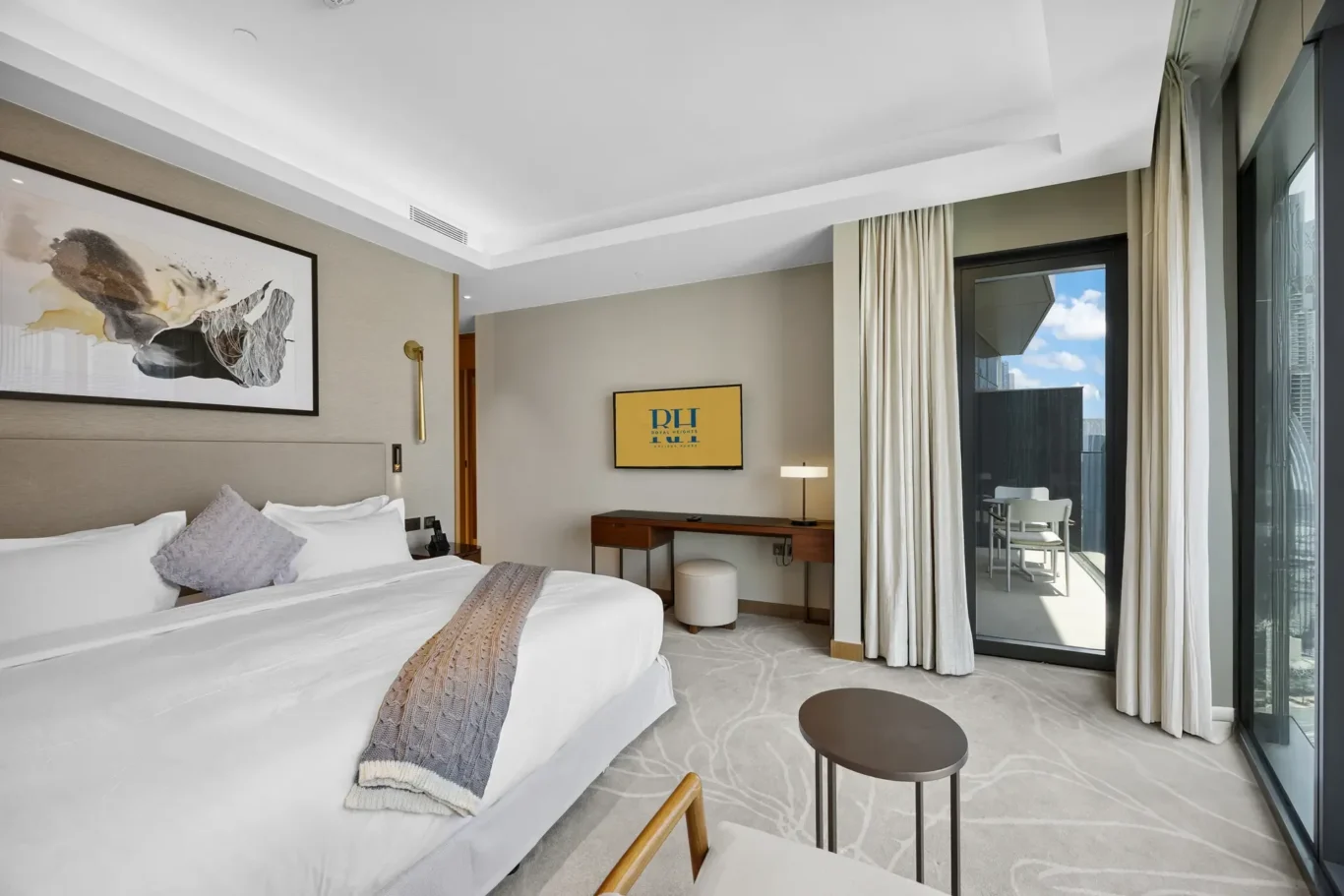 Hotel room with two white beds, beige walls, abstract wall art, and a wooden desk; glass balcony door shows outdoor seating and city view.