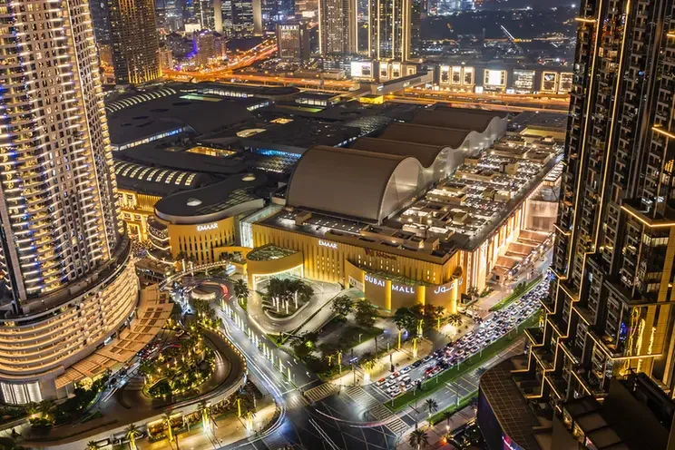 Night aerial view of Dubai Mall and surrounding skyscrapers lit up at night with busy roads below.