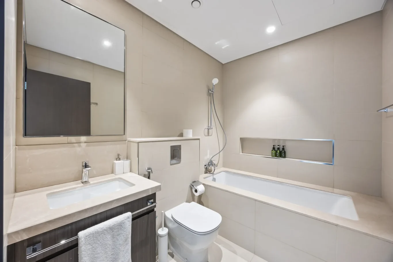 Modern beige-tiled bathroom with a white double vanity, large wall mirror, and chrome fixtures including a handheld shower over a bathtub.