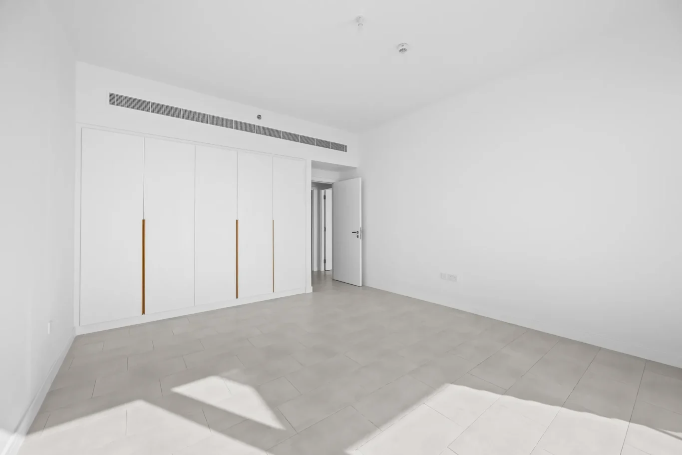 Bright empty room with built-in white wardrobes along the left wall and an open doorway to another space—modern minimalist interior.