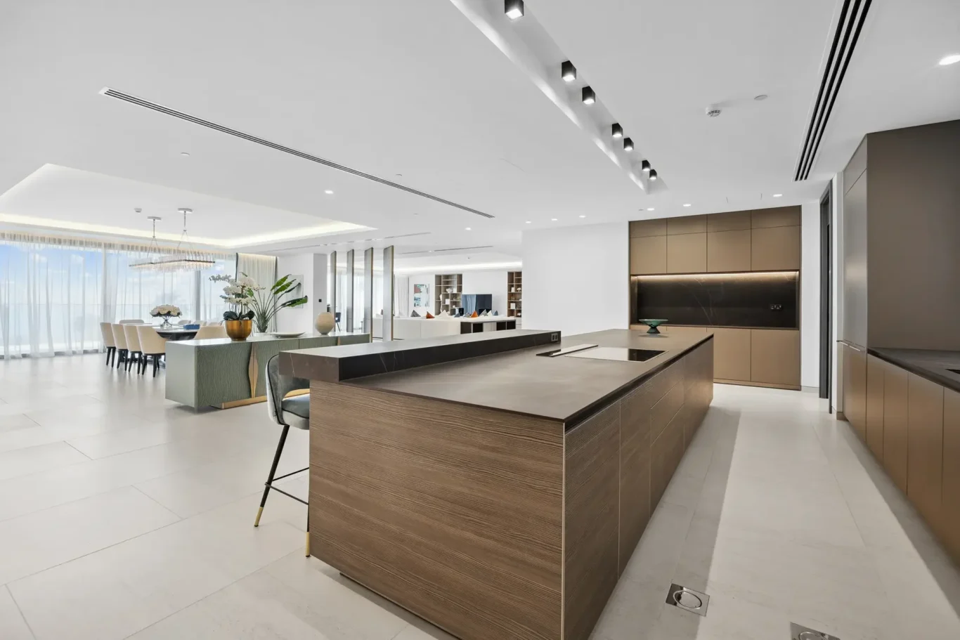 Open-plan kitchen with a large wood-paneled island, beige cabinets, and a bright dining area in the background under soft ceiling lights
