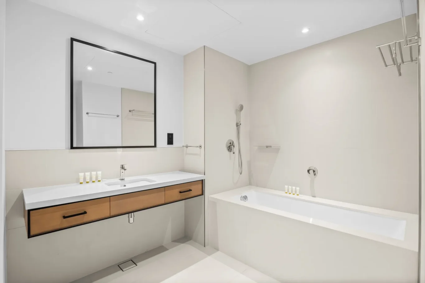 Modern bathroom with white vanity, wooden drawers, large framed mirror, and a bathtub with a wall-mounted shower area.