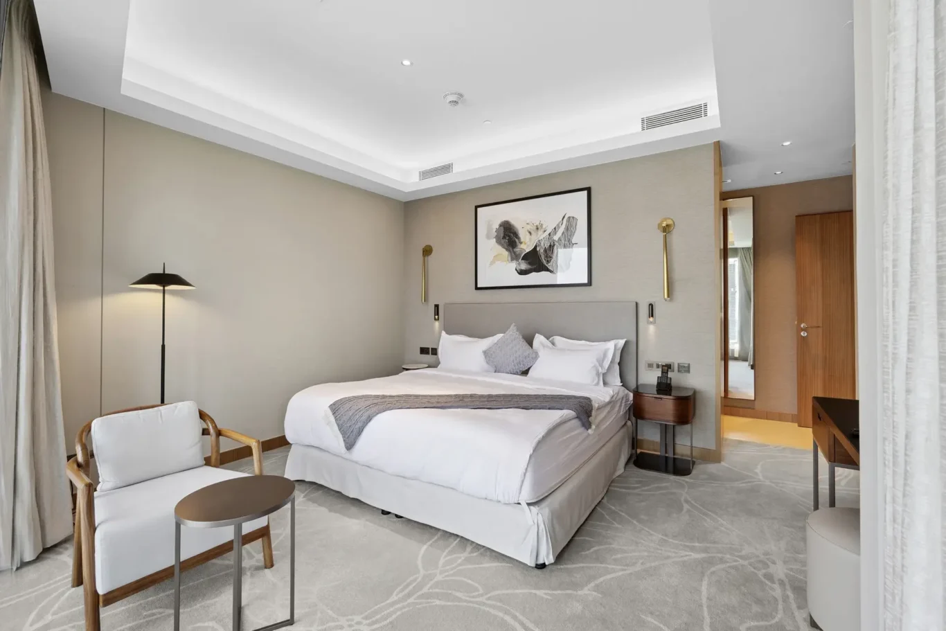 Hotel room with a king bed, gray upholstered headboard, abstract framed art, two nightstands, and a seating area by the window.
