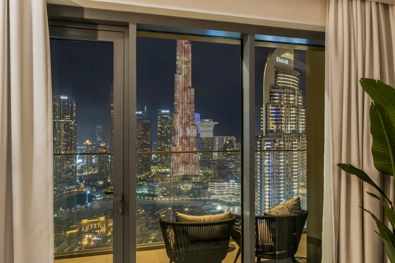 Night city skyline view from a high-rise hotel room balcony with illuminated skyscrapers and glass doors framing the scene, plus wicker chairs in the foreground.