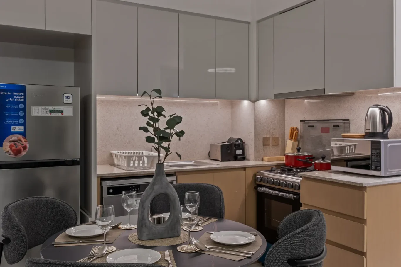Round dining table set for four in a modern kitchen with gray upholstered chairs, a gray vase centerpiece, and place settings for a meal; stainless fridge in the background behind the table portion of the counter.