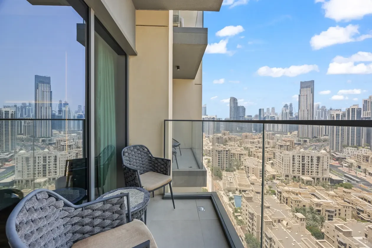 Balcony seating area with woven chairs and glass railing, overlooking a high-rise city skyline on a sunny day.