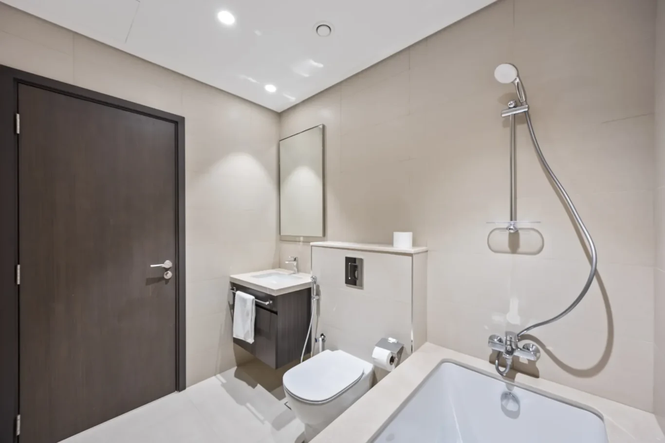 Modern bathroom with wall-mounted vanity, mirror, toilet, and bathtub; wood door and beige tiled walls.