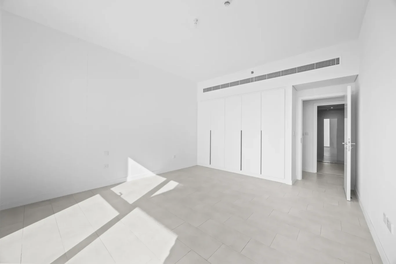 Bright, empty modern room with a long wall of white built-in closets and an open doorway to a hallway, sunlit tiled floor